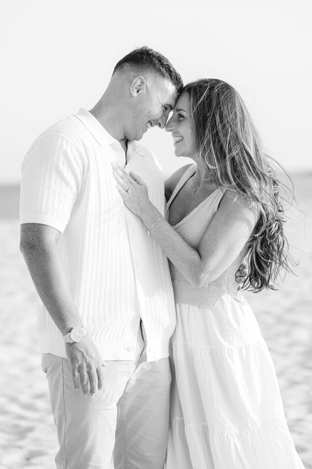 A romantic black and white photo of a couple in white outfits embracing on the beach.