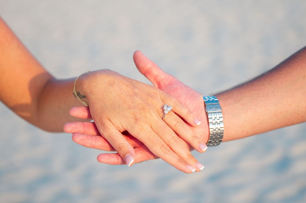 Close-up of hands with engagement ring and bracelet against blurred beach background.