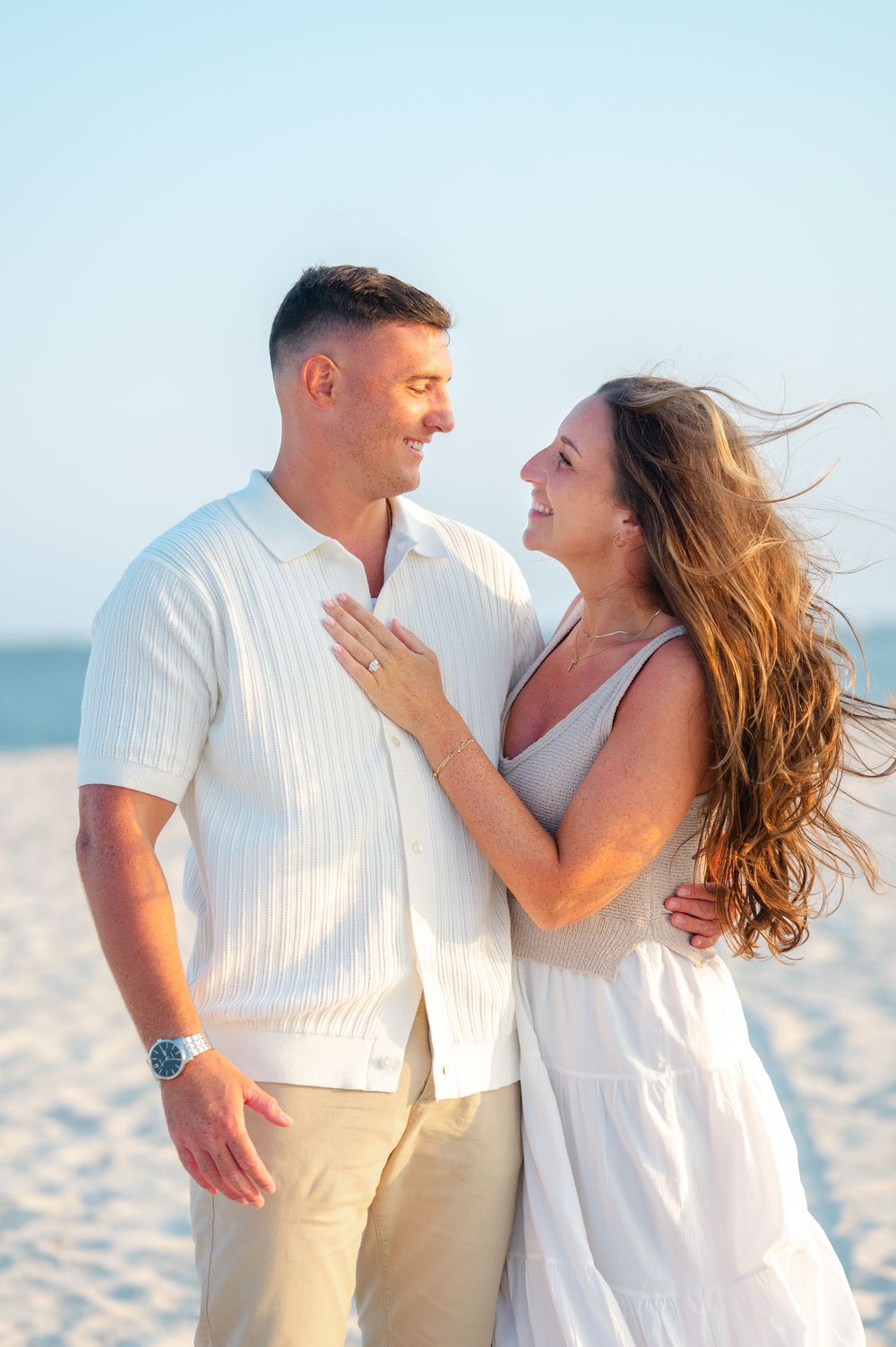 A romantic couple in white outfits shares tender moments while walking on a sunny beach.