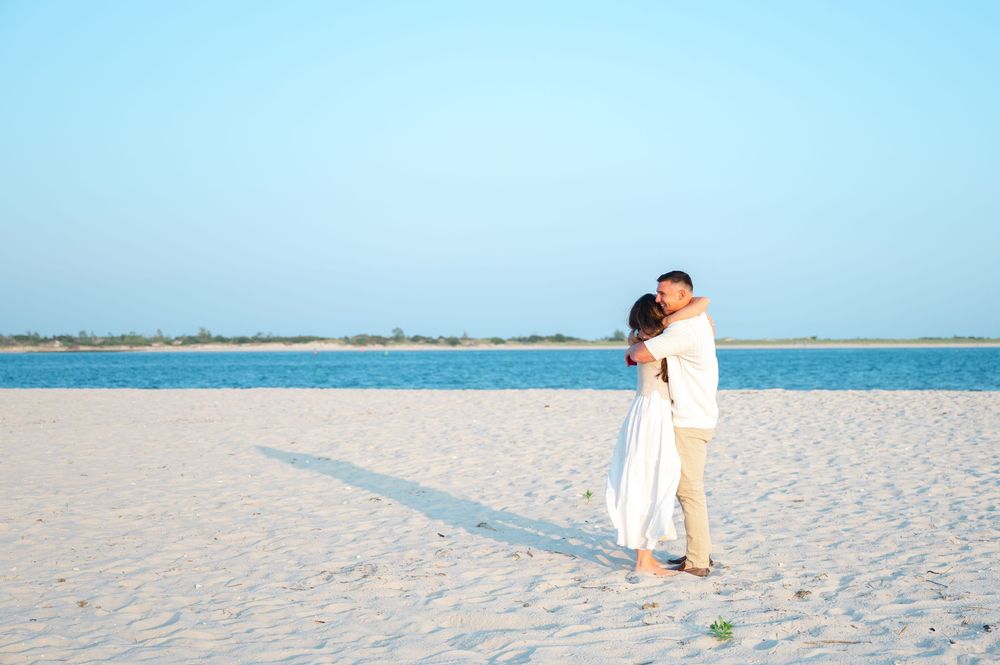 Two people in white outfits embrace on a pristine white sand beach with blue ocean waters in the background.
