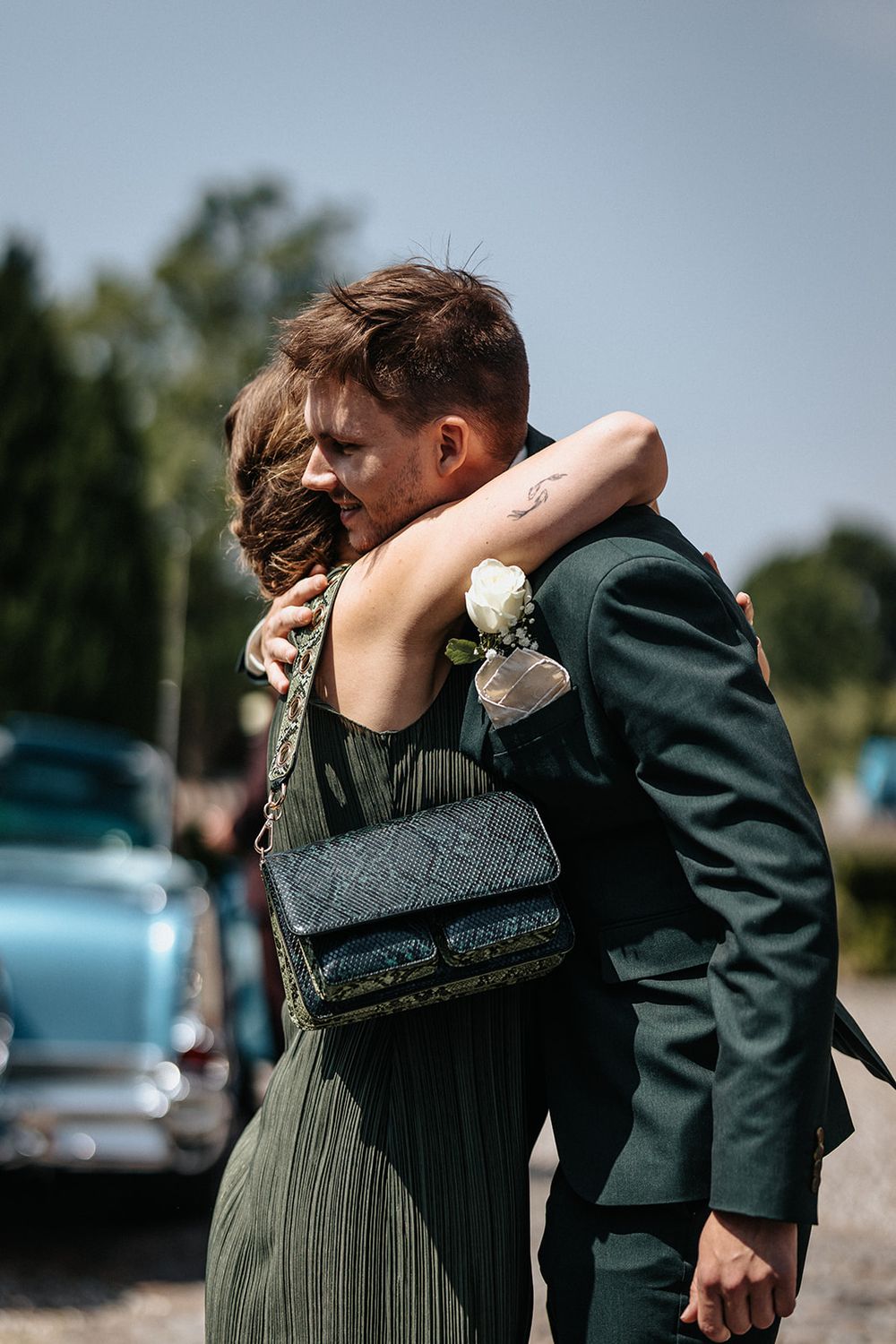 Two people share an emotional embrace outdoors on a sunny day with a green dress and dark suit visible.