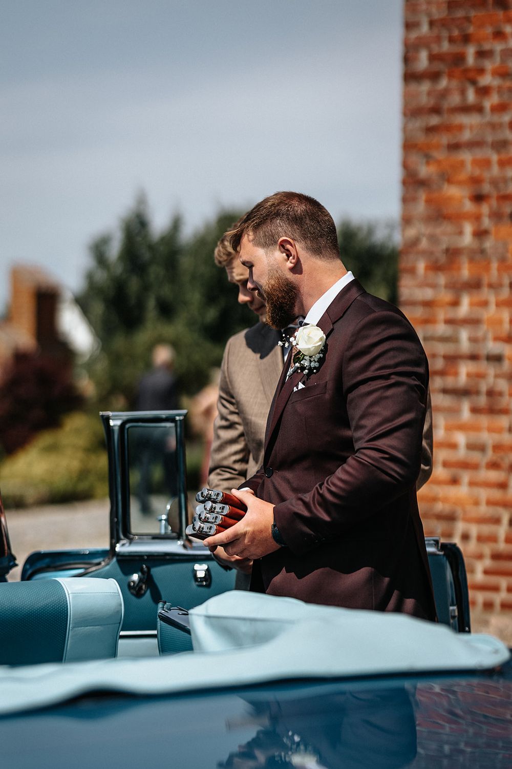 Two people in formal attire stand near a vintage car during an outdoor wedding celebration against a brick wall.