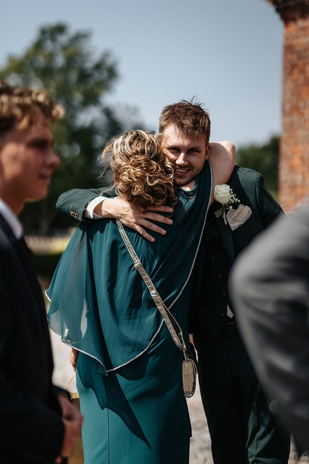 A group wearing formal attire embraces outdoors on a sunny day against a brick wall backdrop.