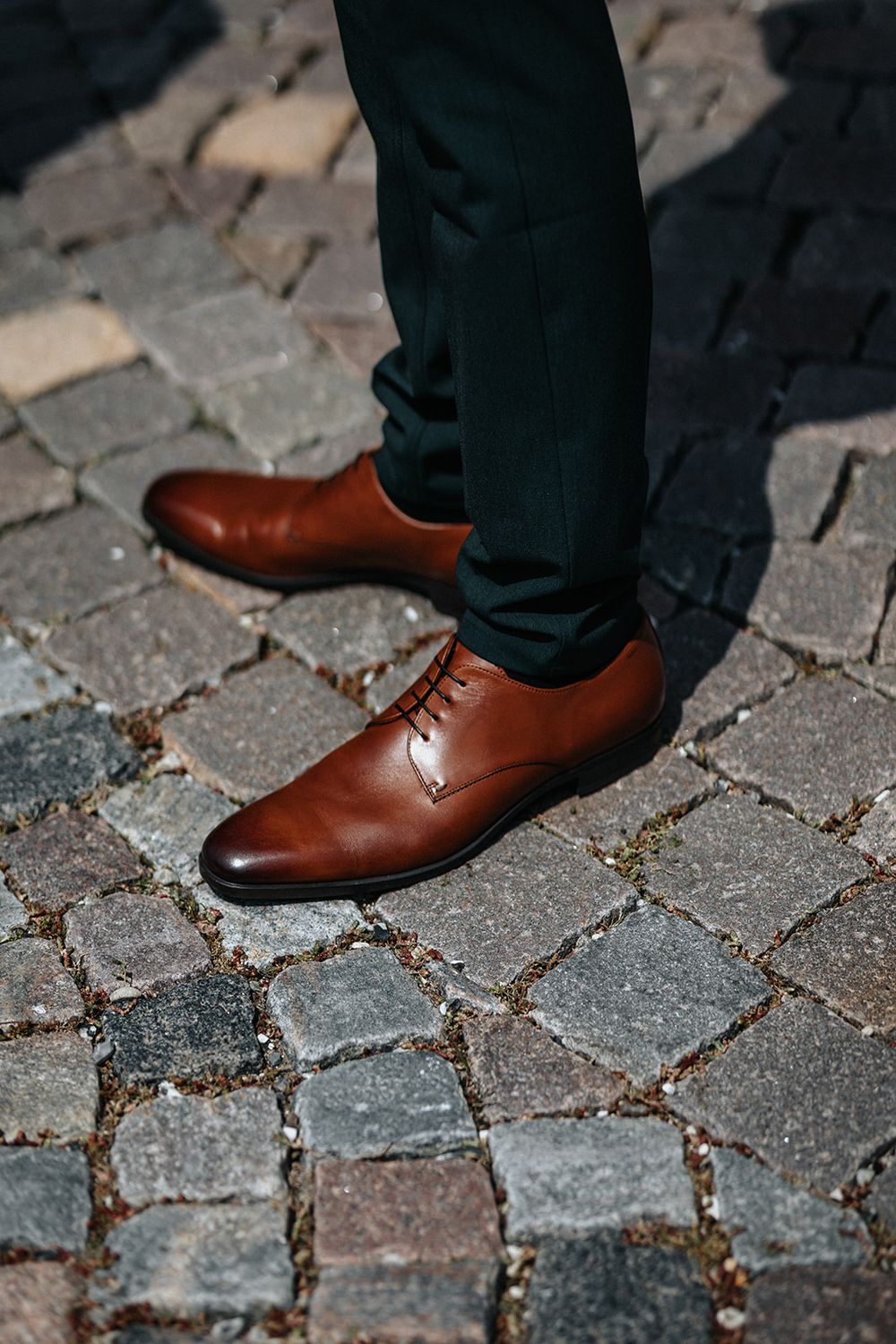 Brown leather dress shoes paired with dark trousers on cobblestone pavement.
