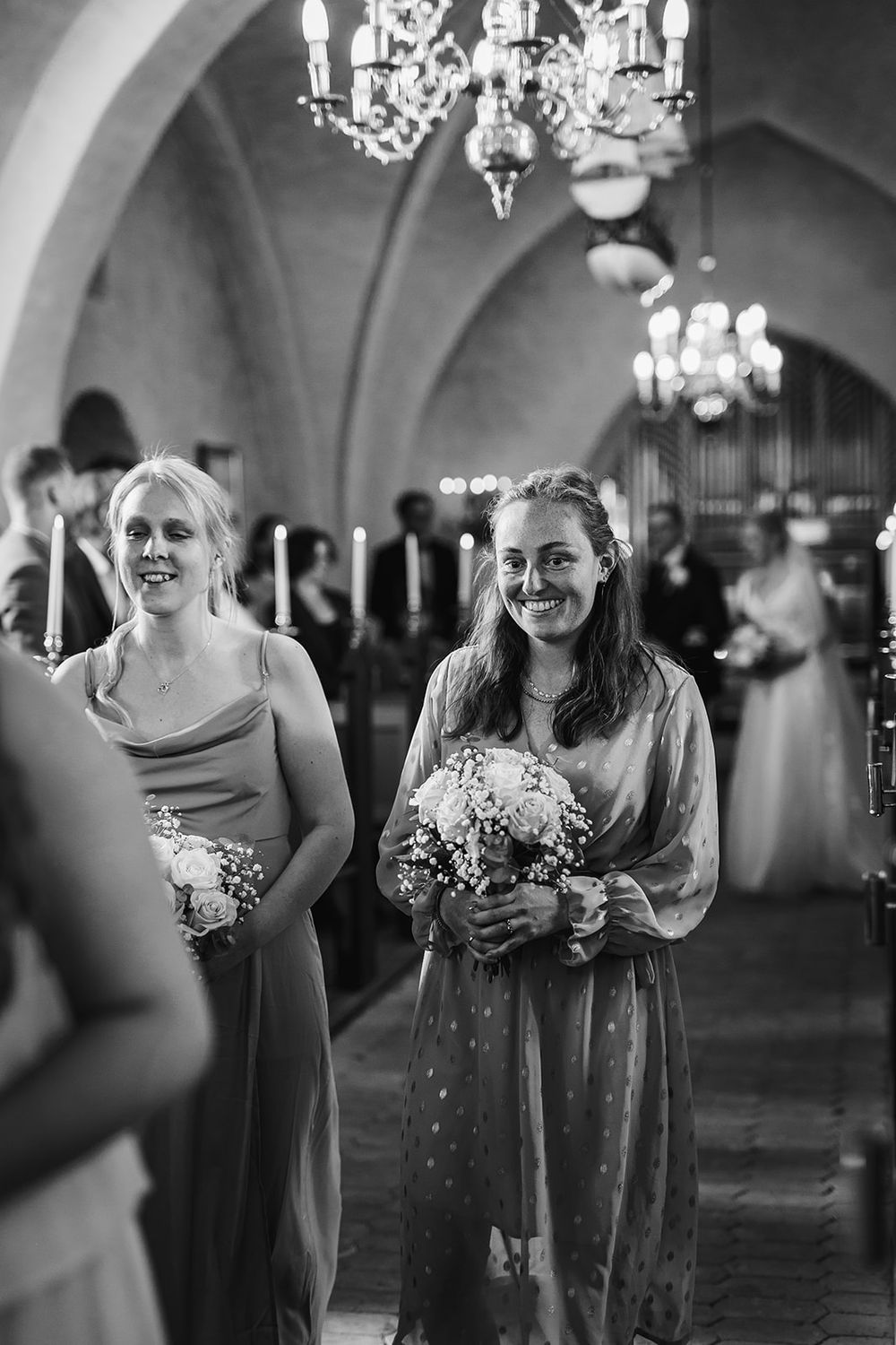 Bridesmaids in elegant dresses share joyful laughter in an ornate church hallway with chandeliers.