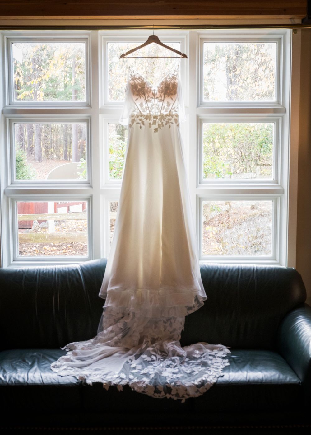 A white wedding dress hangs elegantly by a window, displaying intricate lace details and a flowing train.