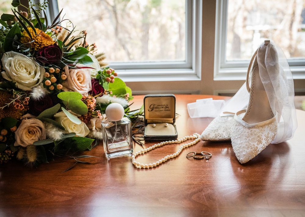 Wedding details including a floral bouquet, white lace shoes, jewelry, and perfume arranged on a wooden surface near a window.