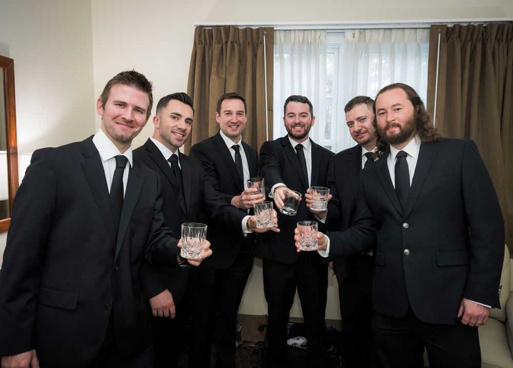 A group wearing formal black suits and ties stand together raising glasses in celebration at an indoor event.