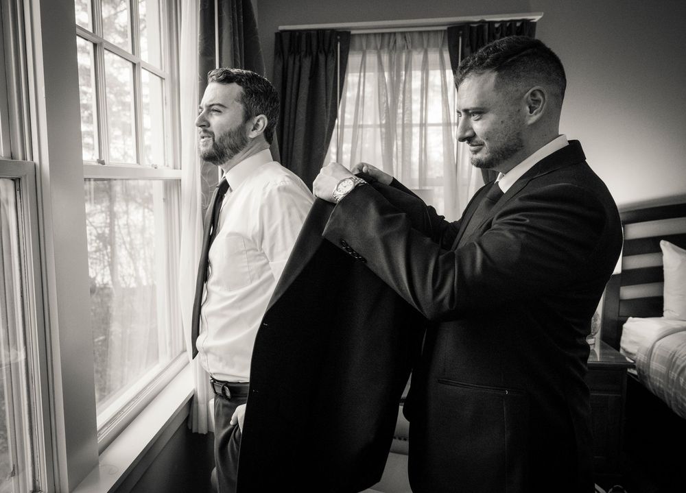 Several men in formal black suits adjusting clothing and preparing near a bright window.