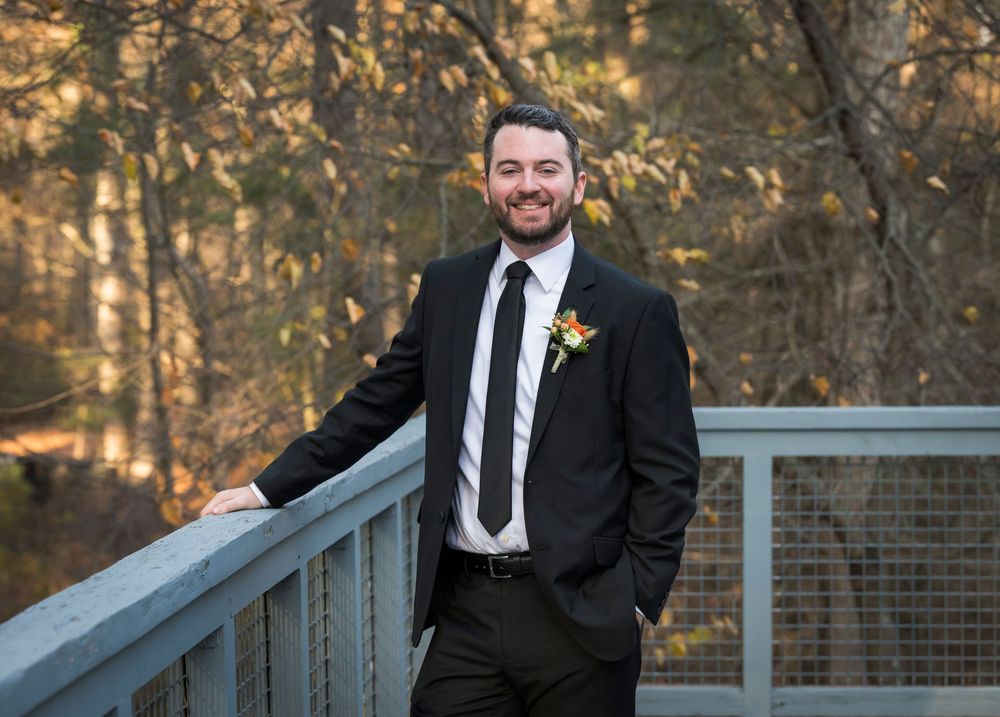 A person in a black suit and tie smiles while standing against an autumn forest backdrop and railing.