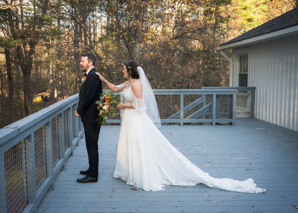 A bride in a white gown with long train stands with groom in black suit on a wooden deck during autumn.