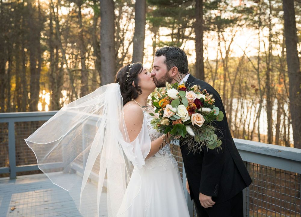A romantic wedding couple embraces at sunset in a wooded setting with a flowing veil catching the golden light.