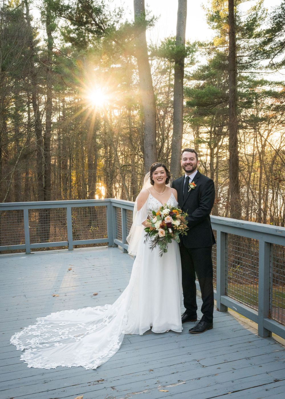 Wedding couple poses on wooden deck surrounded by pine trees at sunset with dramatic sunburst through branches.