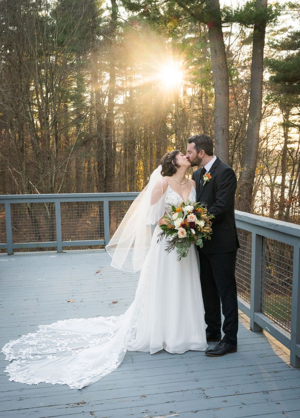 A couple shares a romantic moment on a wooden deck at sunset with sunlight streaming through pine trees.