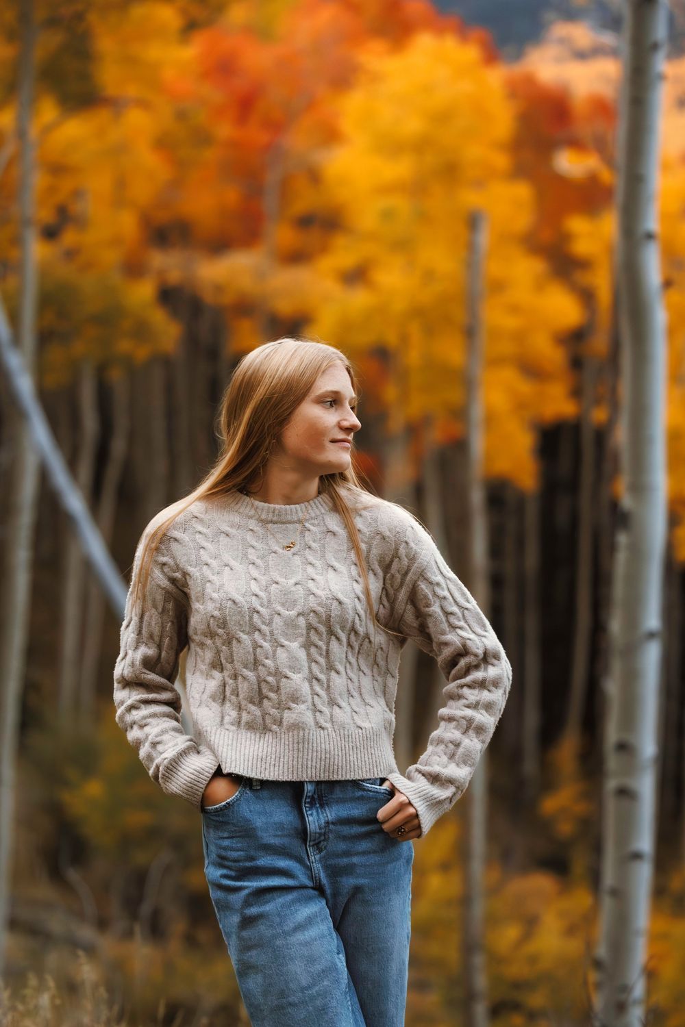 Young blonde woman in cream cable-knit sweater and jeans poses among colorful fall aspen trees.