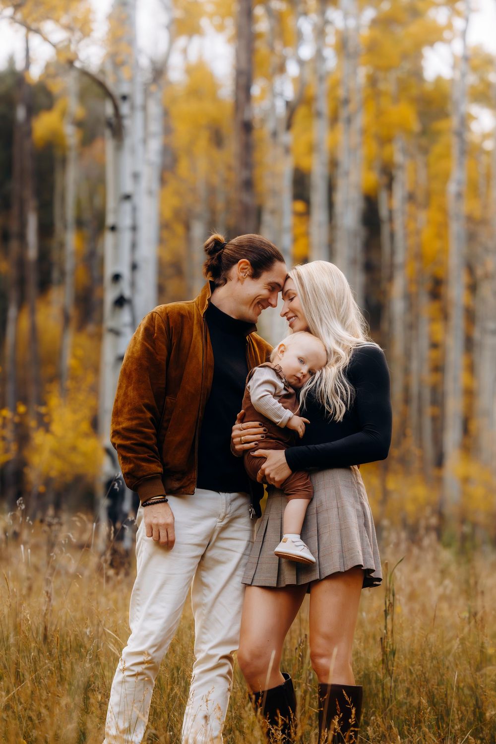 Young couple tenderly touches foreheads while holding their baby in a golden autumn aspen grove.