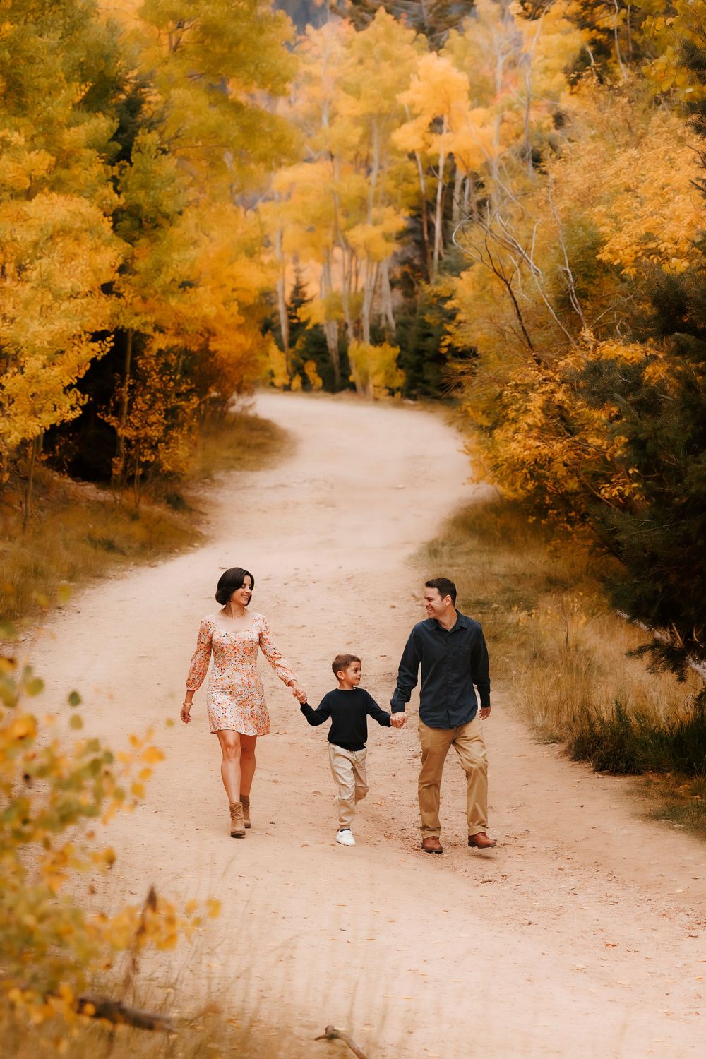 Family of three walking hand in hand down a winding dirt path through golden autumn trees.