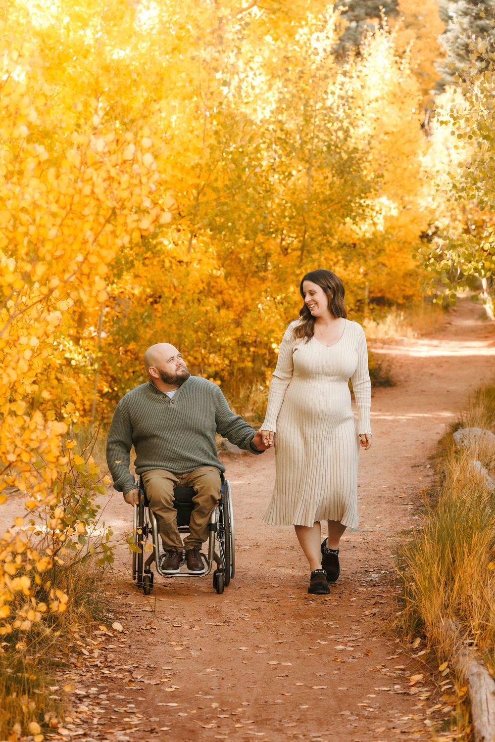 Pregnant woman holds hands with husband in wheelchair on a scenic path lined with golden fall trees.