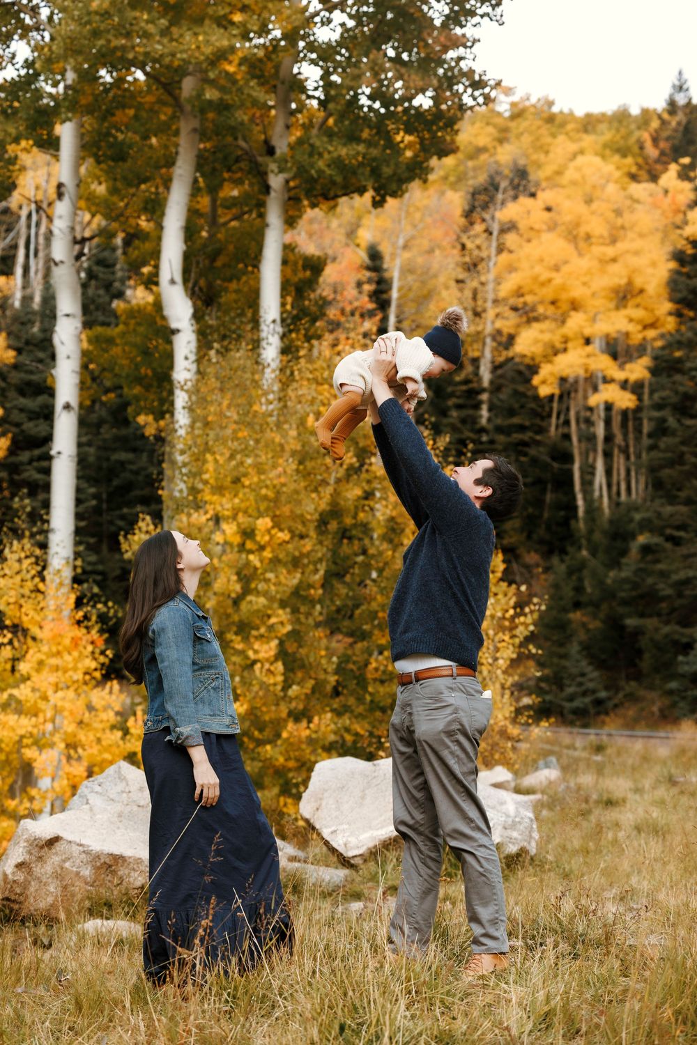 Father tossing baby in the air while mother watches among golden autumn aspen trees.