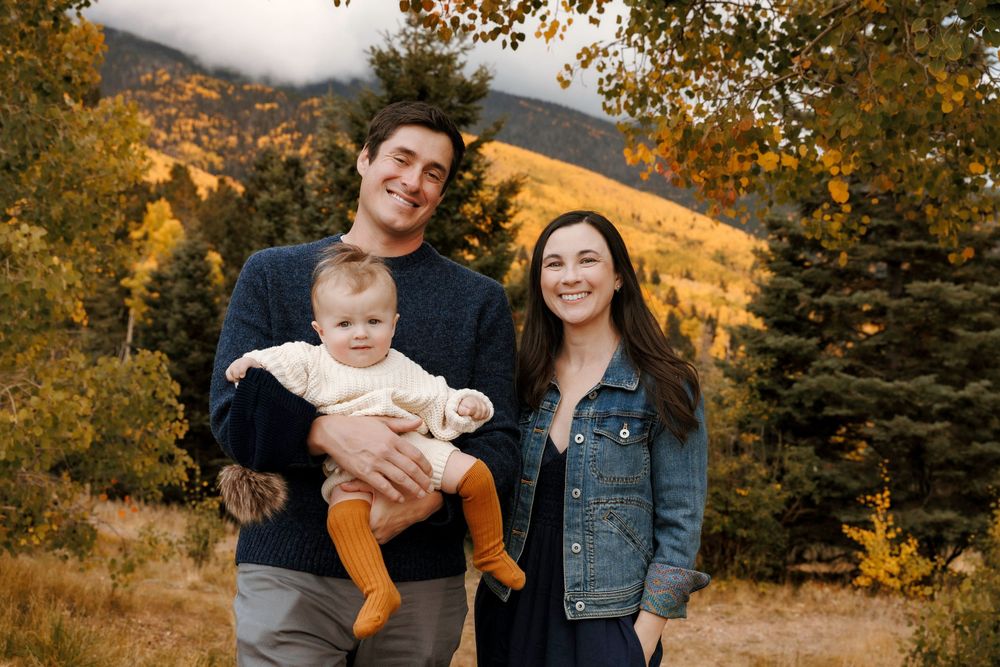 Smiling family of three holding baby outdoors with golden mountain foliage in background.