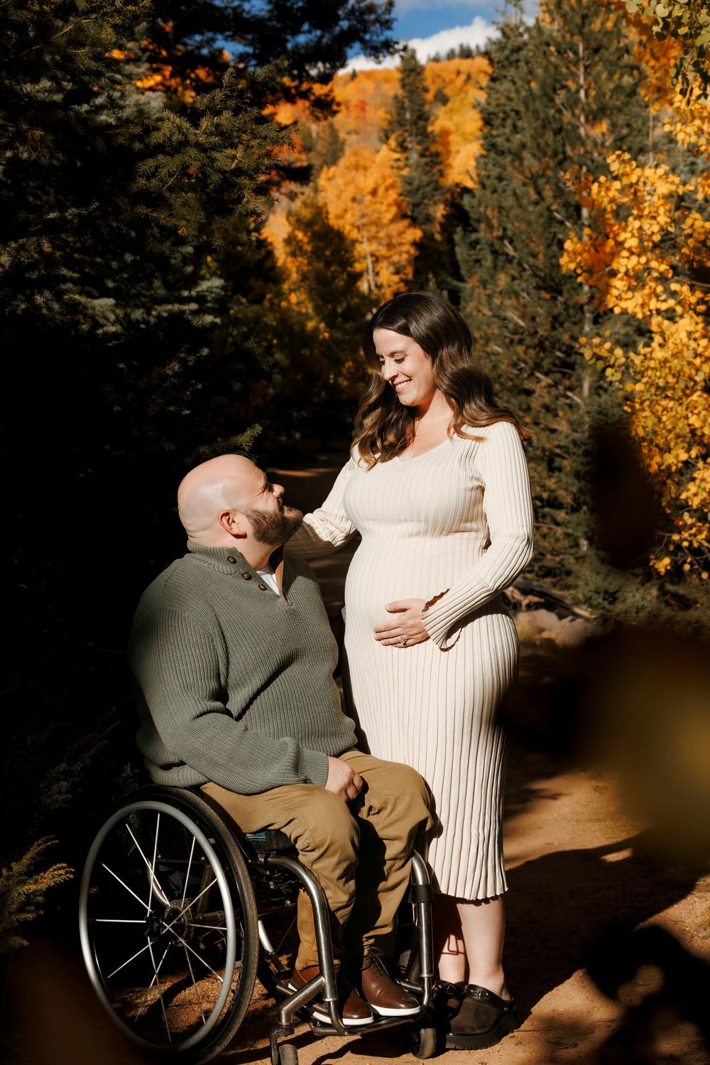 Pregnant woman in cream dress with man in wheelchair sharing tender moment among autumn foliage.