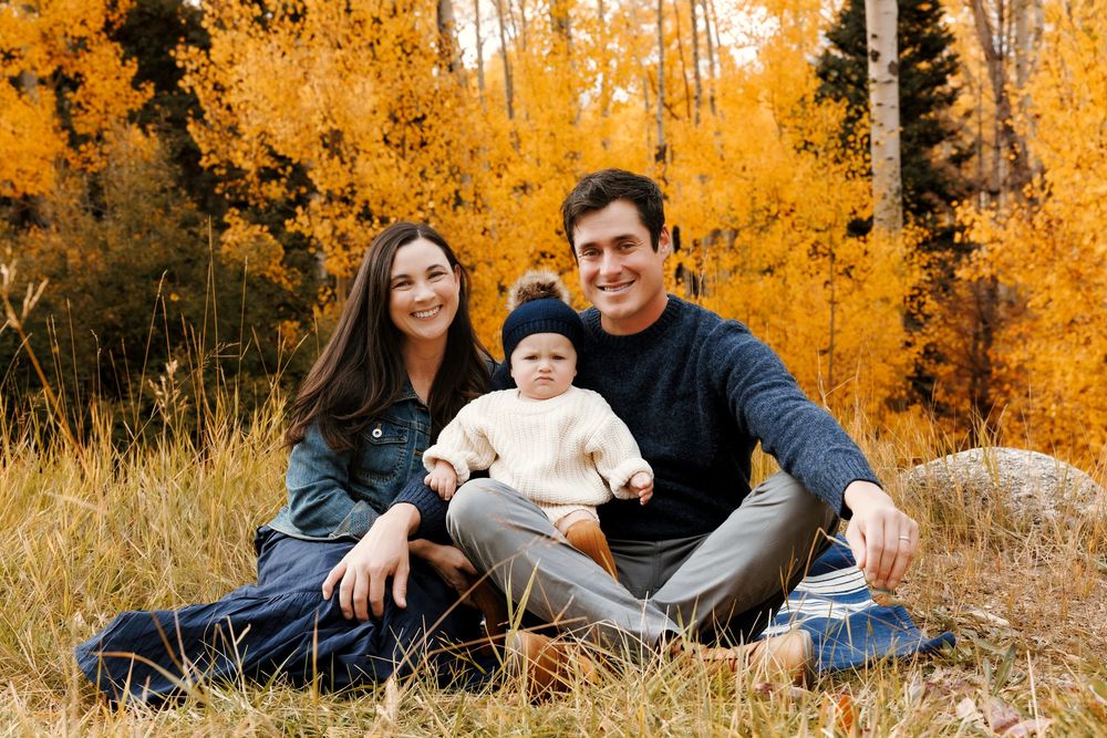 Smiling family of three with baby sitting on a blanket among golden fall aspen trees.