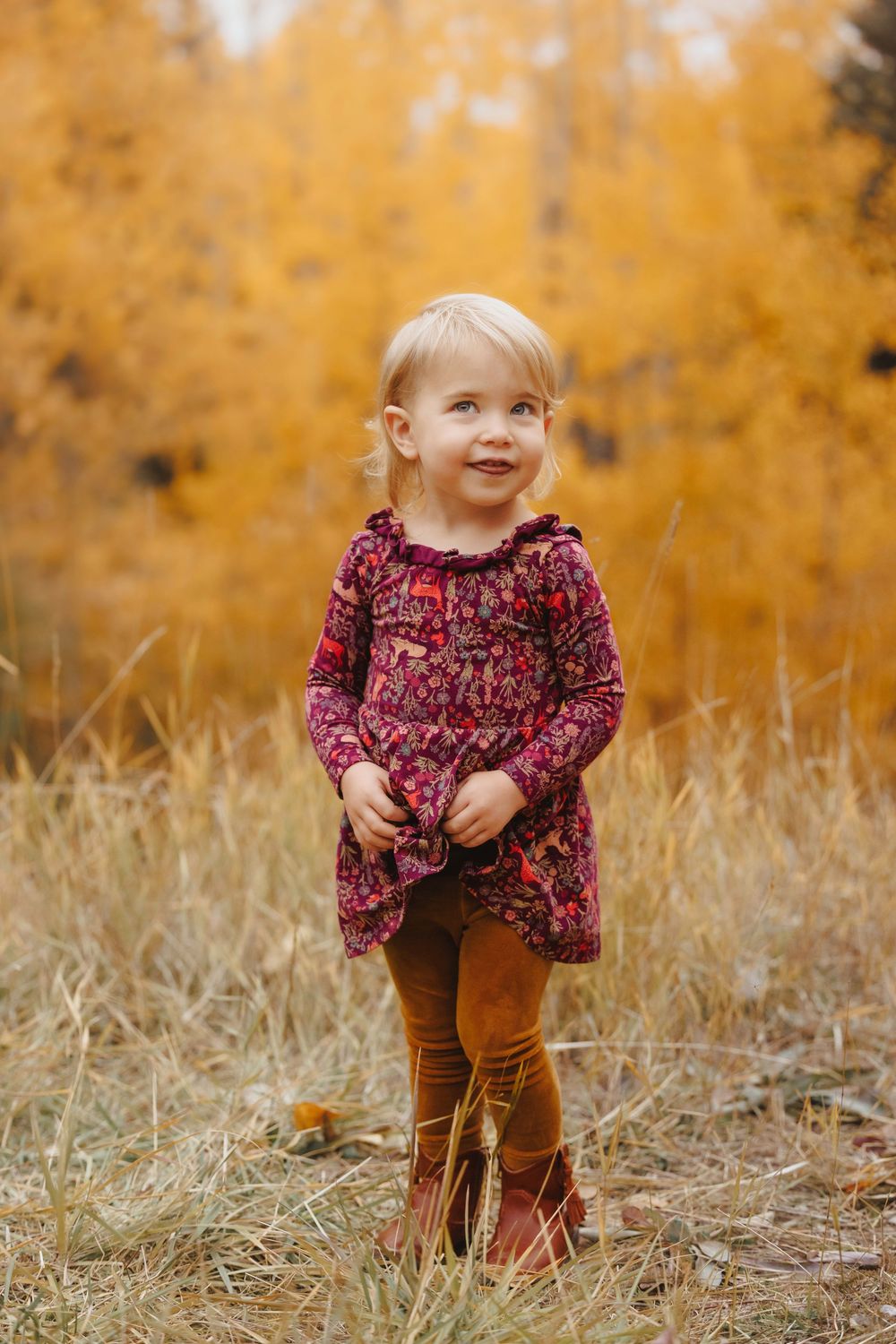 Adorable toddler girl in floral dress and brown boots stands in a field of golden autumn foliage.