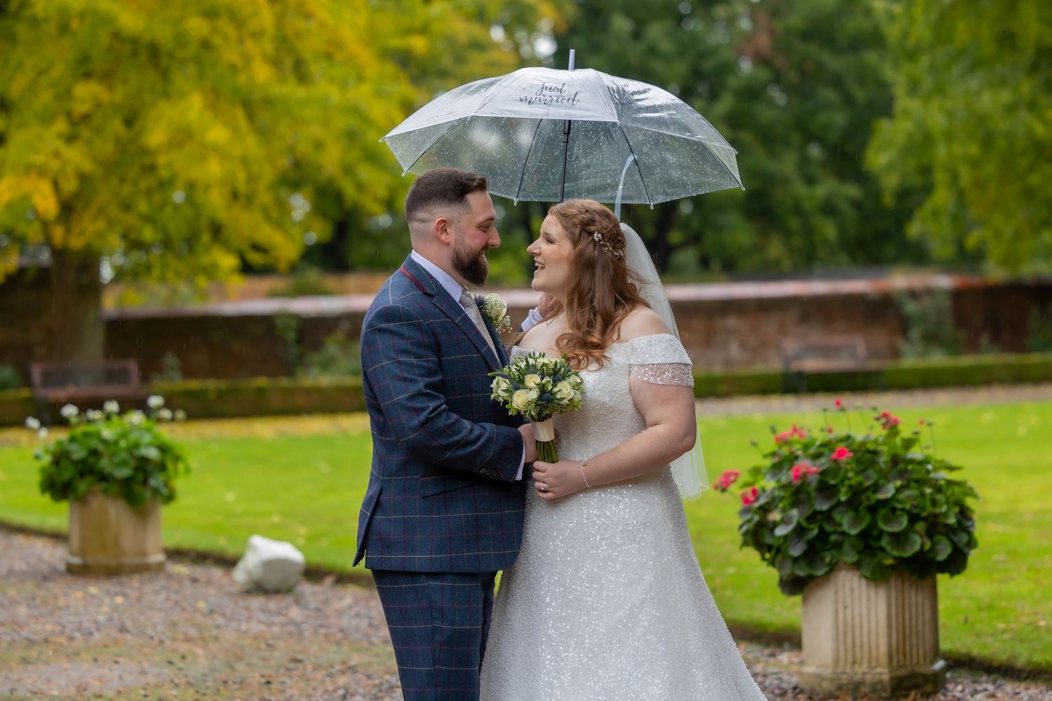 A couple in wedding attire shares a romantic moment in a lush garden setting with autumn foliage in the background. Eshott Hall.
