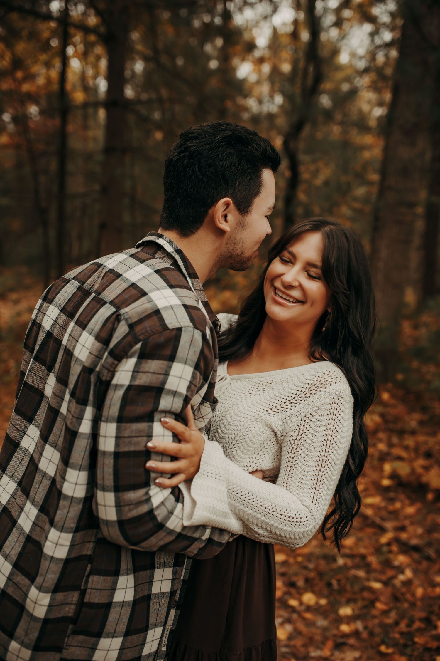 A couple embraces in a romantic autumn forest setting, wearing cozy sweaters among fallen leaves. Creative Art By Bree LLC