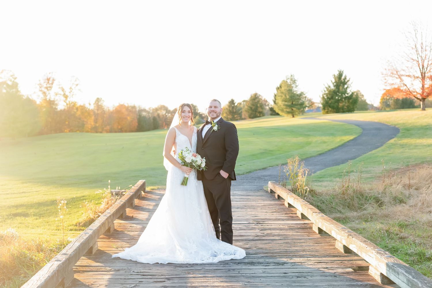 A couple in formal wedding attire stands on a wooden bridge during golden hour sunset.