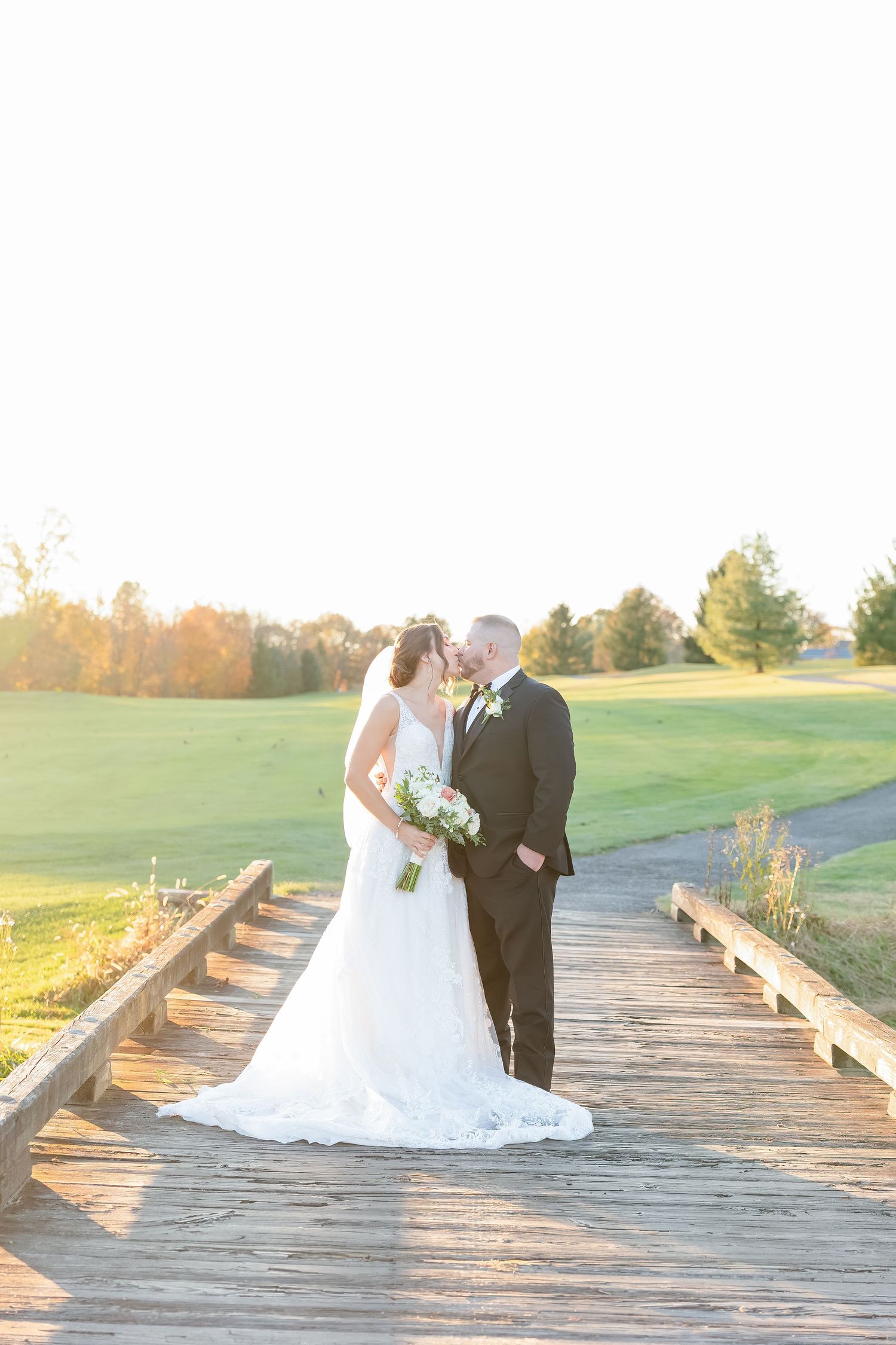 A couple in formal wedding attire poses on a wooden bridge at sunset on a golf course.