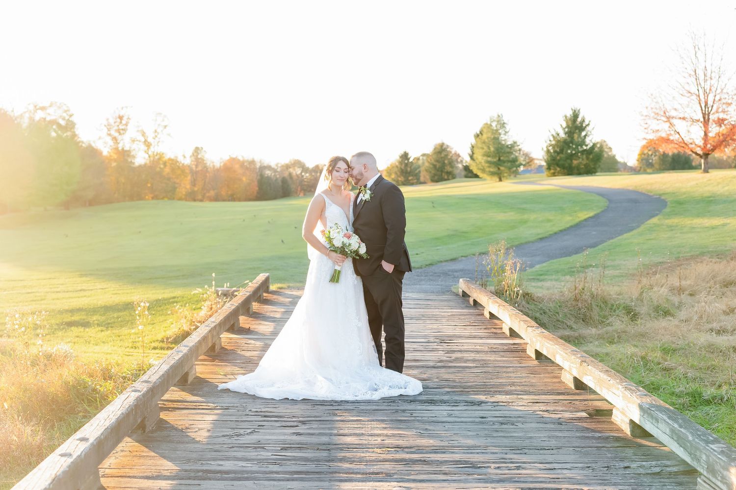 A couple stands on a wooden boardwalk bridge during sunset in a scenic outdoor setting.