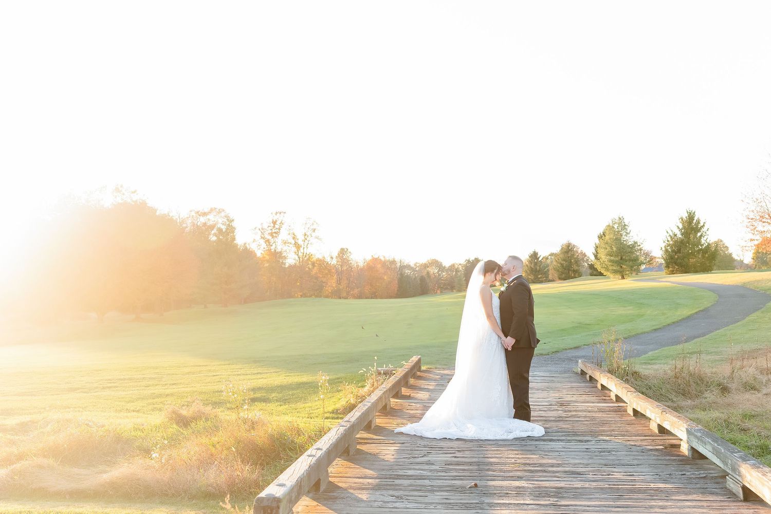 A scenic rural dirt path leads through green fields at sunset with golden sunlight illuminating the landscape.