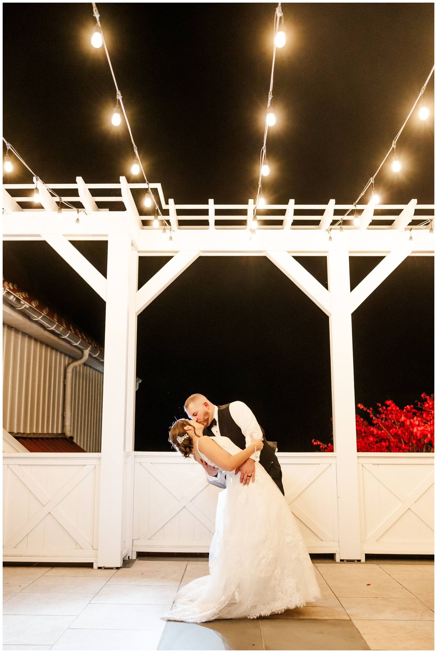 A couple dances under glowing string lights on a white deck with a romantic red floral backdrop at night.