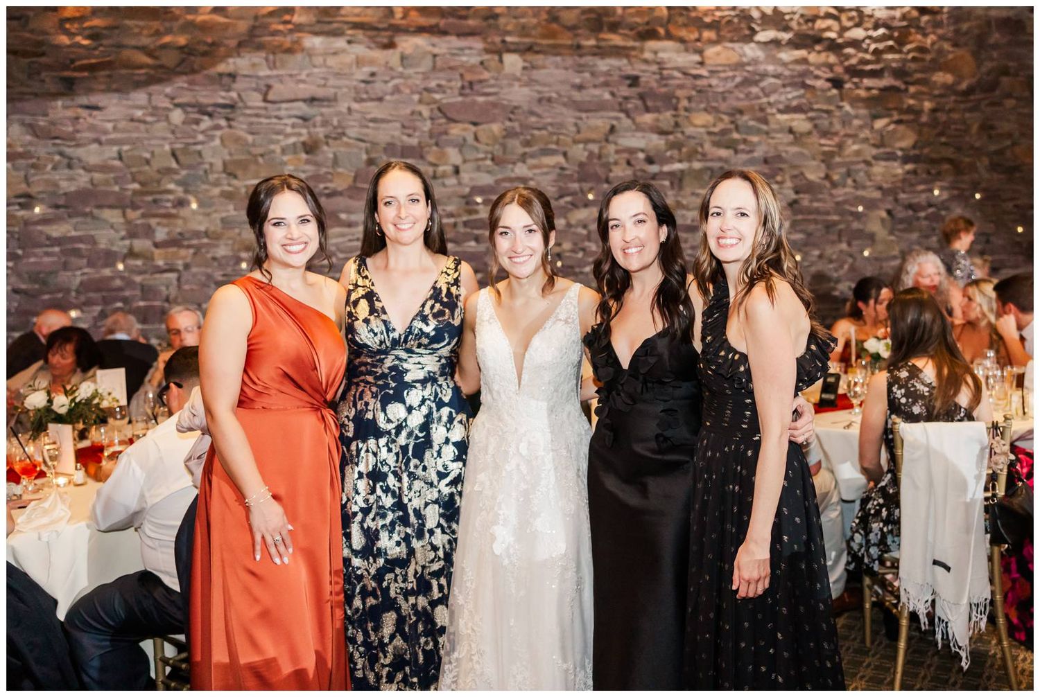 Wedding party members in rust orange and floral dresses pose together against a rustic stone wall at sunset.