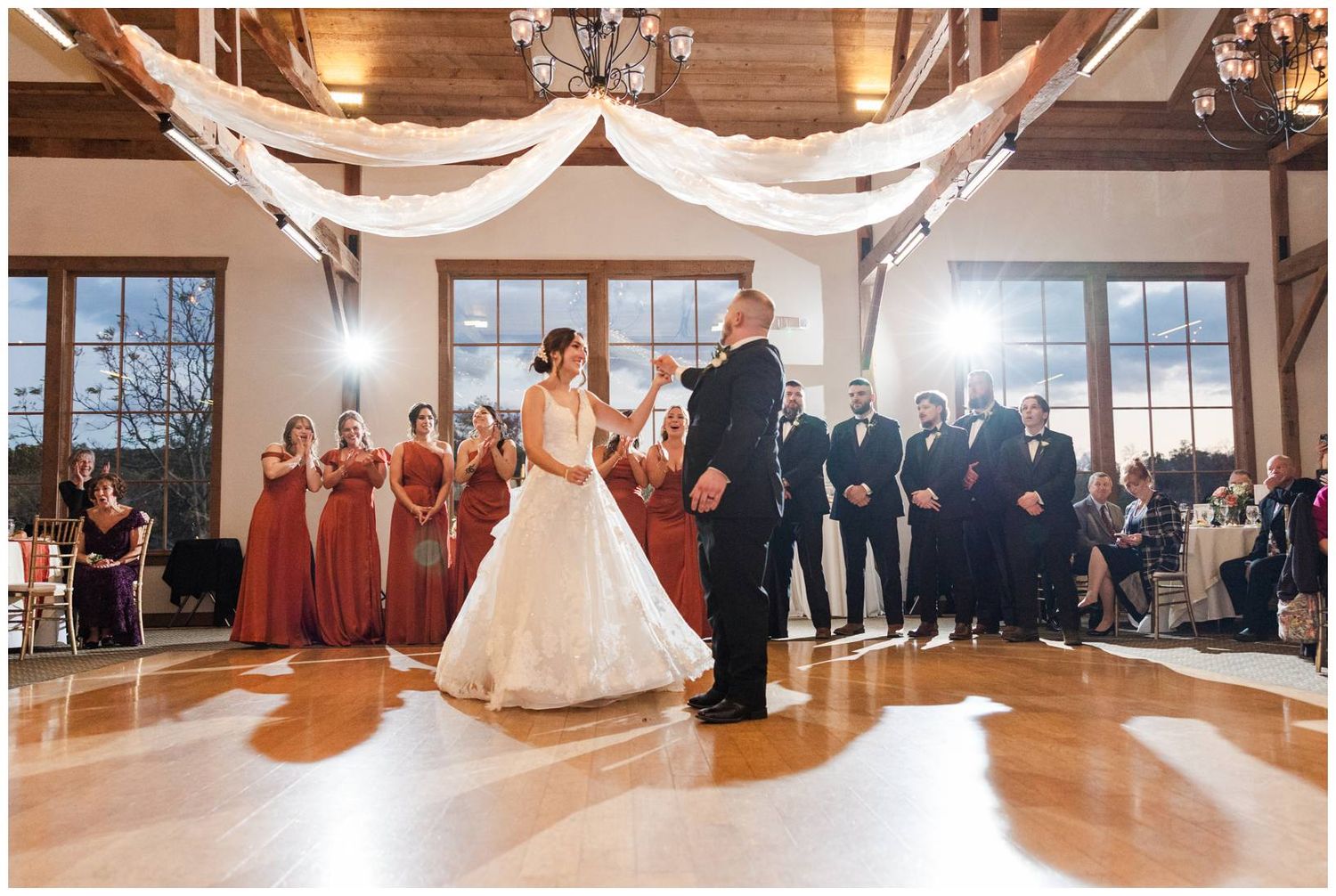 Wedding party members watch a couple sharing their first dance in an elegant rustic venue with large windows.