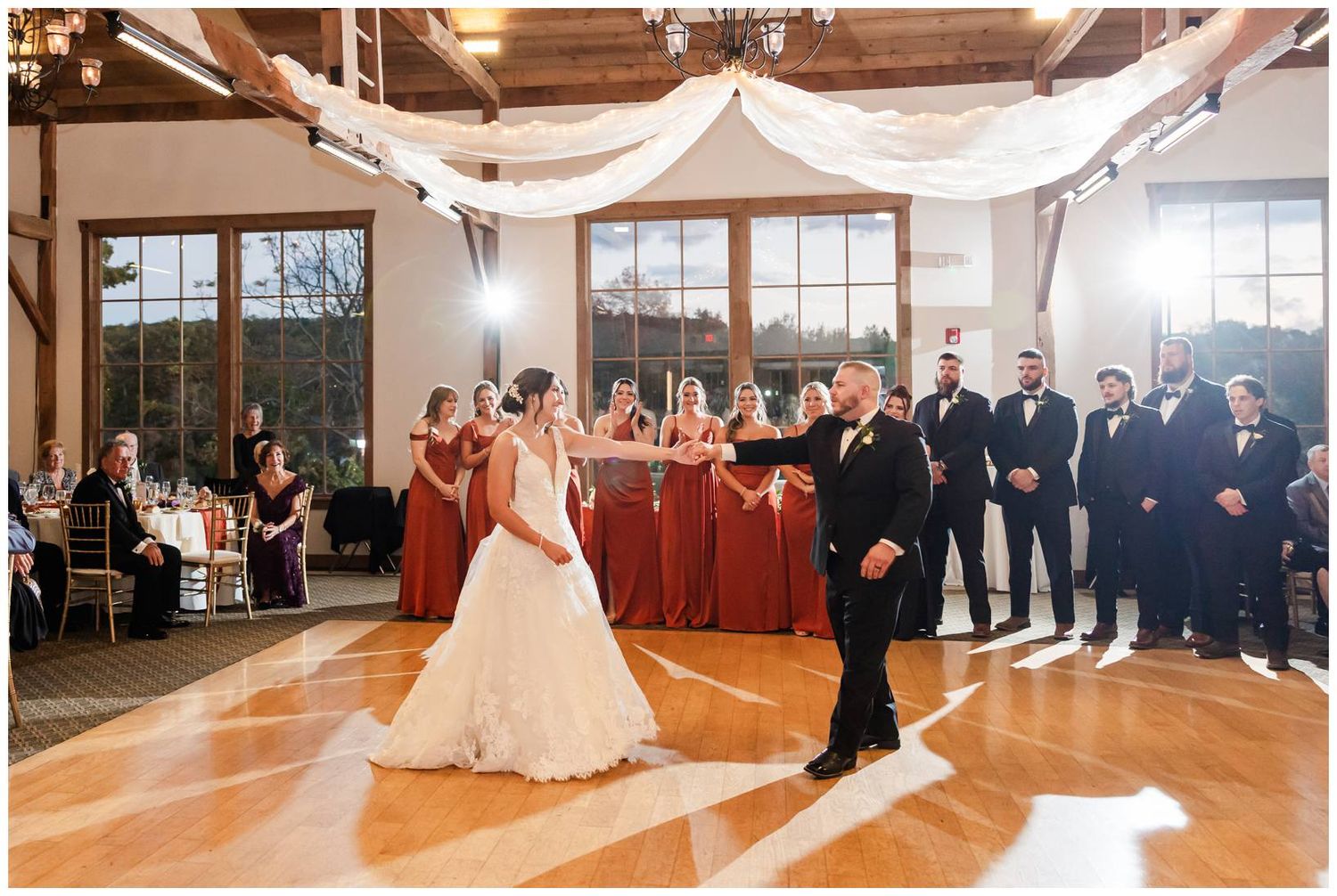 A couple shares their first dance at a wedding reception in an elegant venue with draped fabric and large windows.