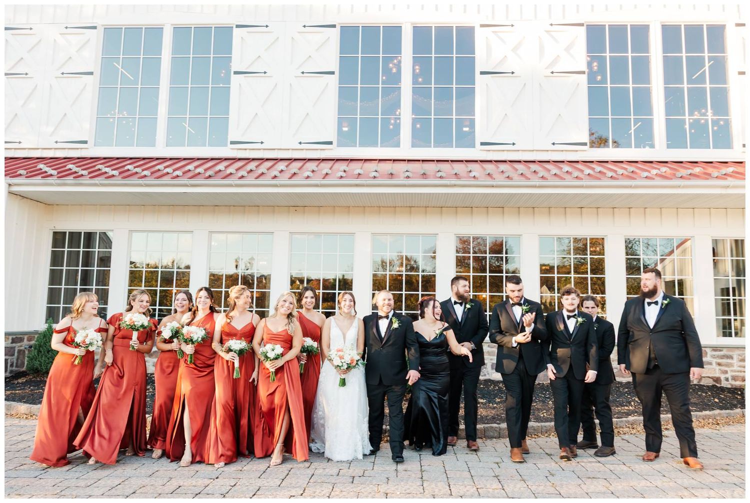 Full wedding party portrait in front of white venue with bridesmaids in red and groomsmen in black.