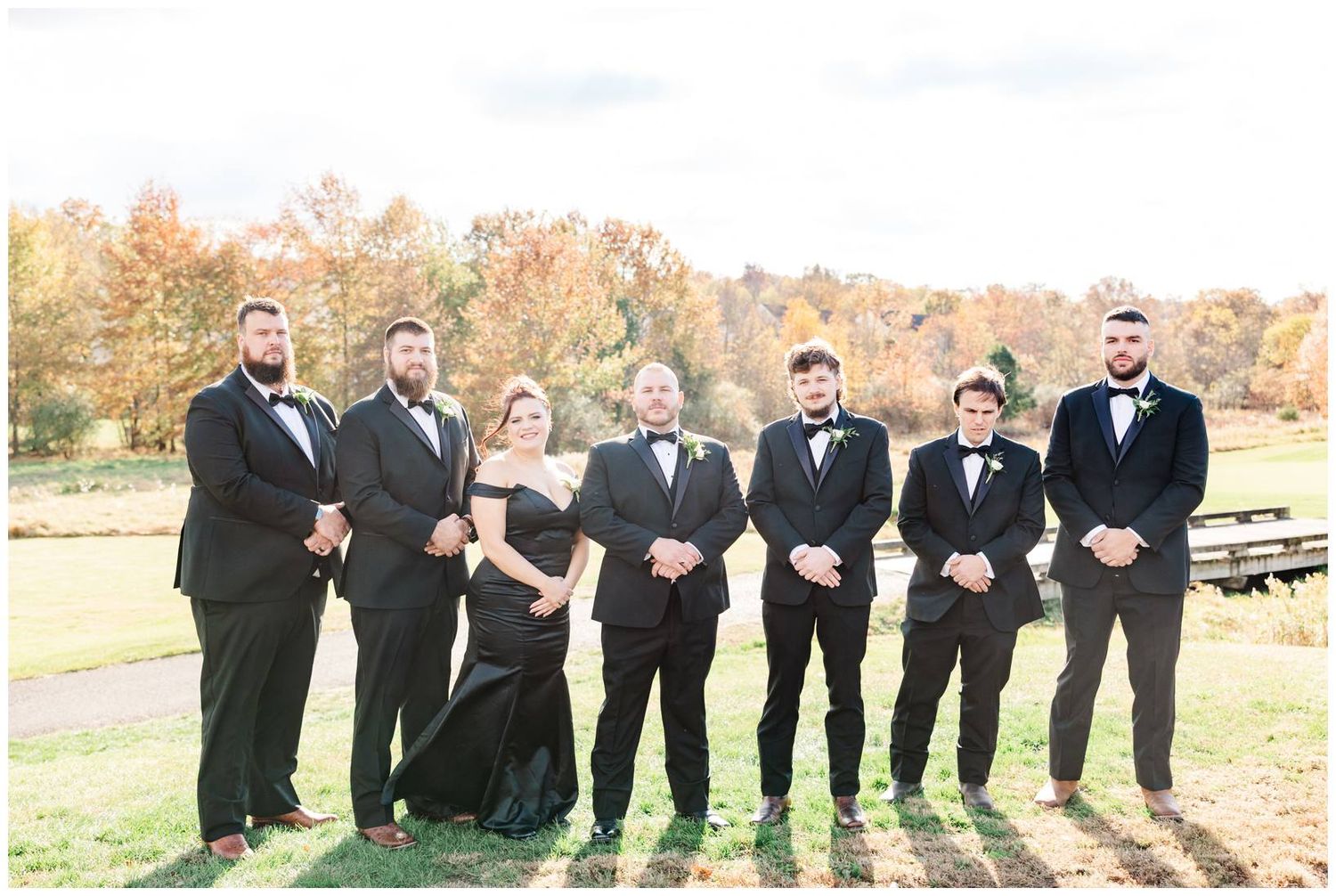Wedding party in black formal attire poses together on a grassy field during autumn with fall foliage in background.