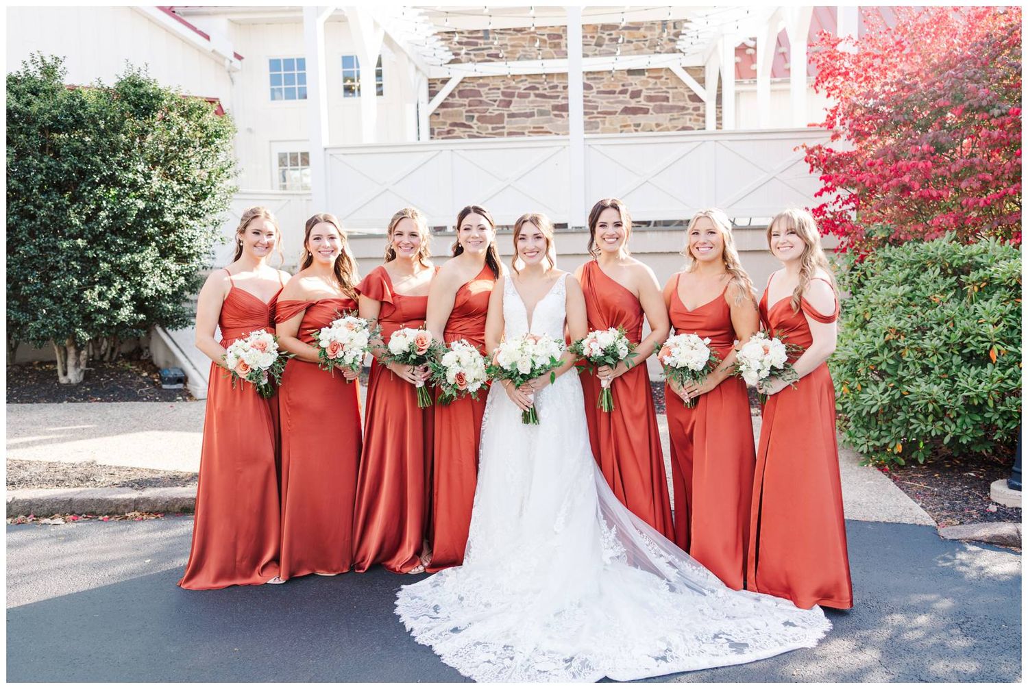 Wedding party wearing red bridesmaid dresses and white bridal gown pose outside venue with autumn foliage.