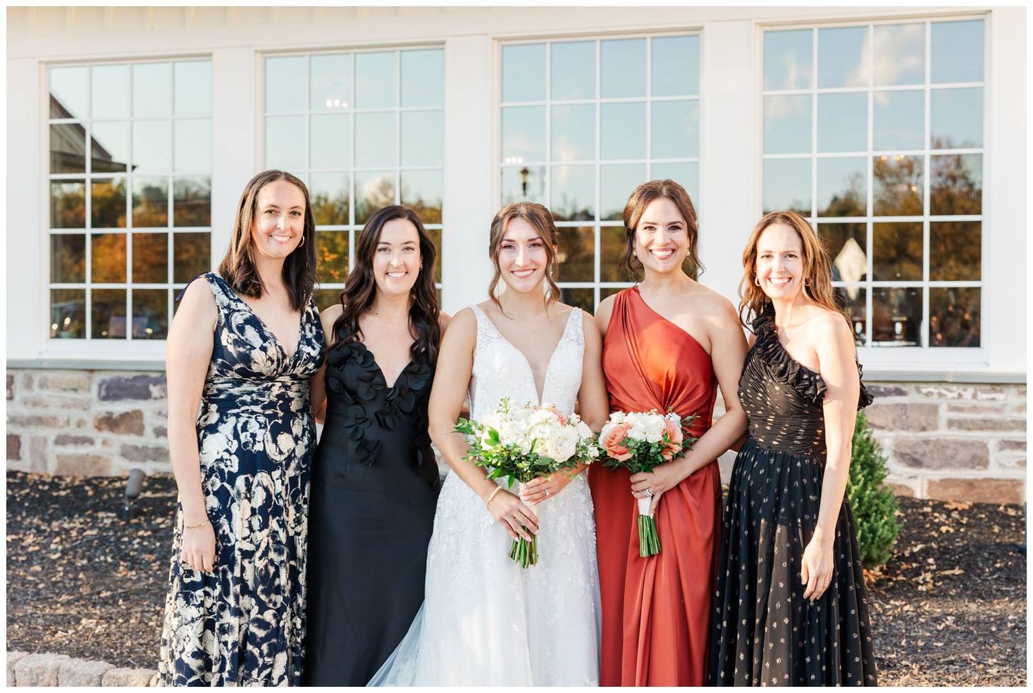 Group photo of wedding party members in formal attire standing together outside a stone building.