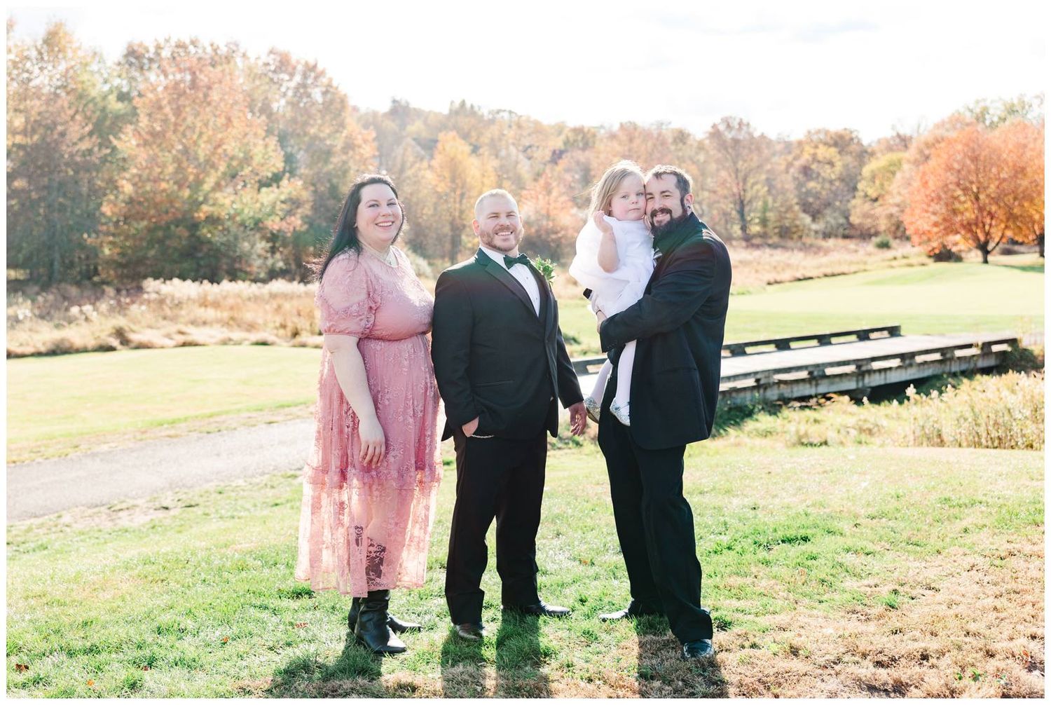 Wedding party members gather outdoors on a grassy hill during autumn with fall foliage in the background.