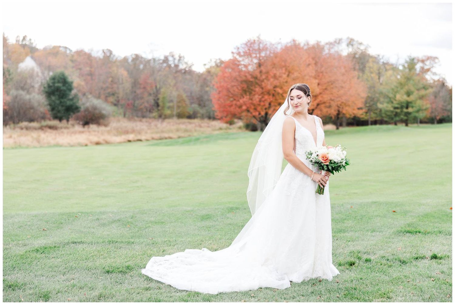 A bride in a flowing white gown and veil poses in a field with vibrant autumn foliage in the background.