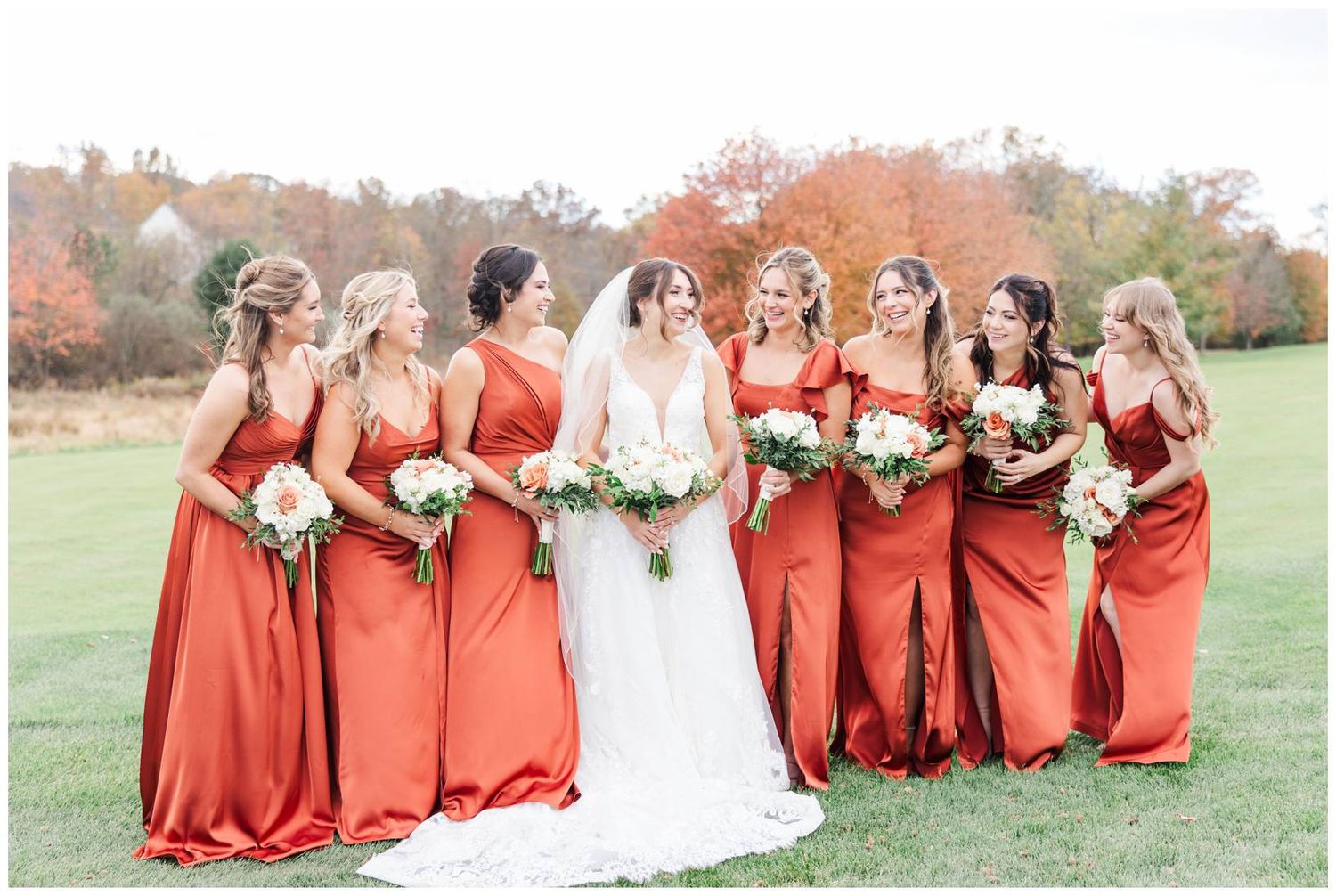 Bridal party wearing vibrant red dresses pose together on grassy lawn during autumn wedding celebration.