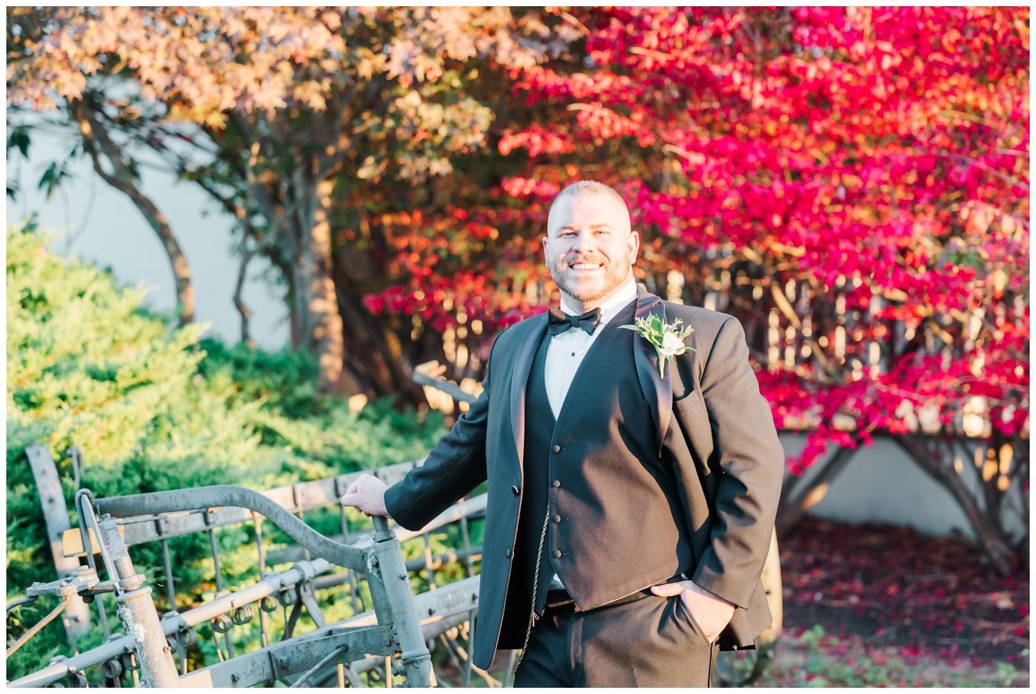 A person in a tuxedo stands near a wooden fence with vibrant red and yellow autumn foliage in the background.