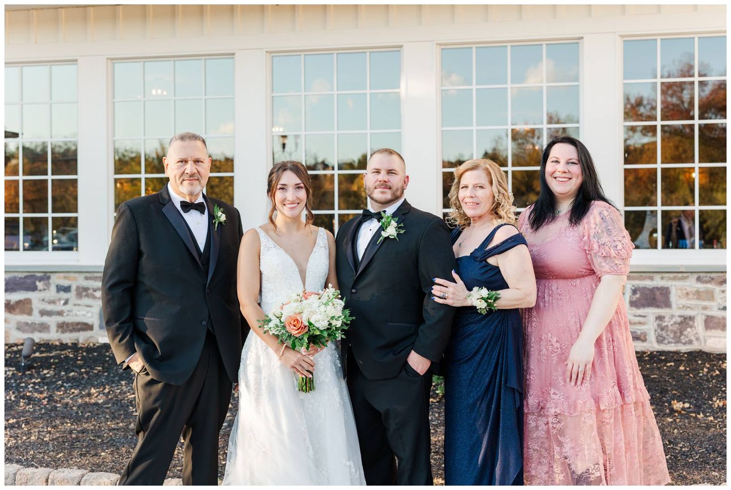 Wedding party poses together outside stone building with large windows during autumn celebration.