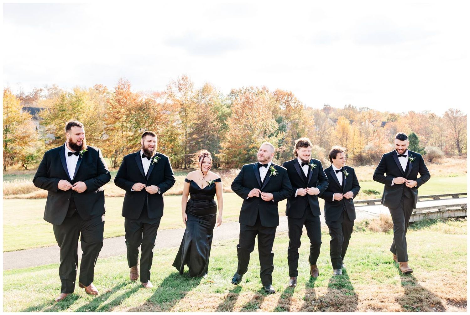 Groomsmen in black tuxedos walk across lawn with autumn trees in background.