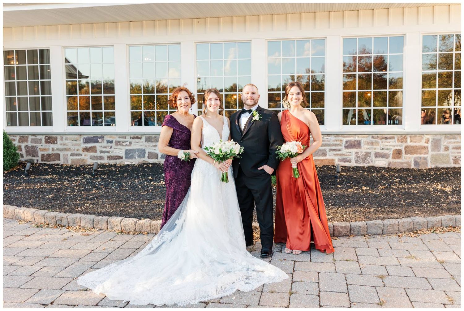 Wedding party members gather outside white venue building with stone accents and paved courtyard.