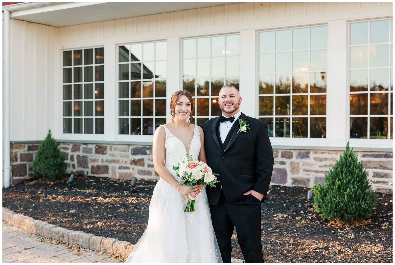 Elegant wedding couple poses outside stone and white venue building with large windows.