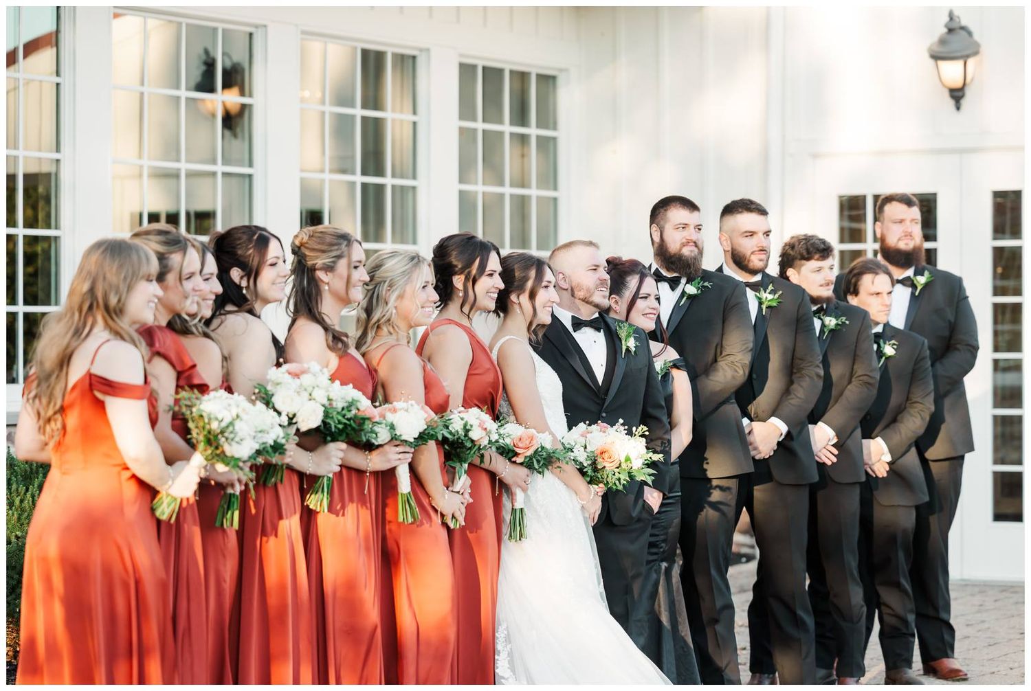 Wedding party in formal attire poses together outside white building with classic architecture.